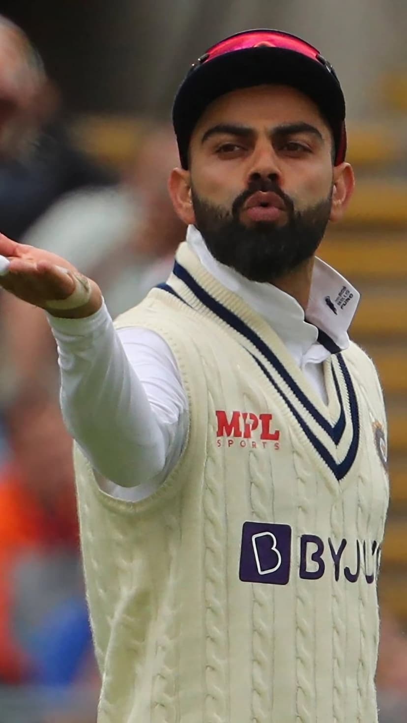 Virat Kohli in a cream cable-knit vest and blue cap gestures during a Test match with a blurred stadium behind him.