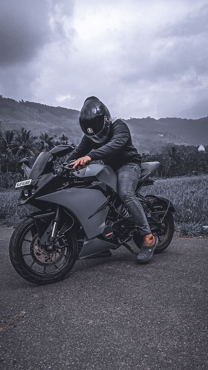 Motorcyclist on a sportbike sits on an asphalt road with palm trees and a cloudy sky in a grayscale landscape.