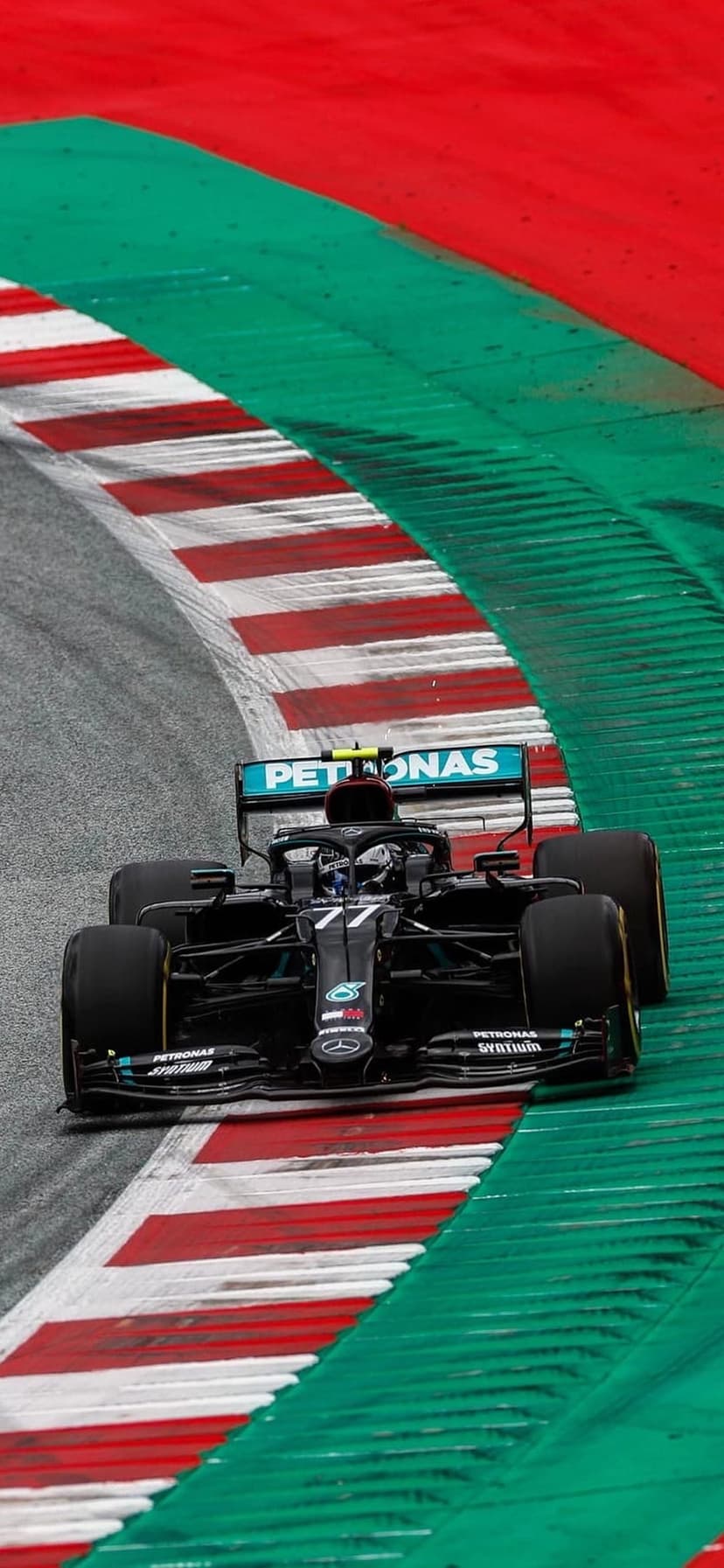 Mercedes Formula 1 car with number 77 leans into a track corner with red and white kerbs and green run-off area.