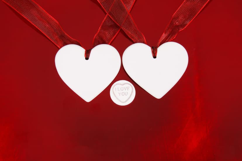 White ceramic hearts suspended by red satin ribbons against a scarlet background with a silver circular charm.