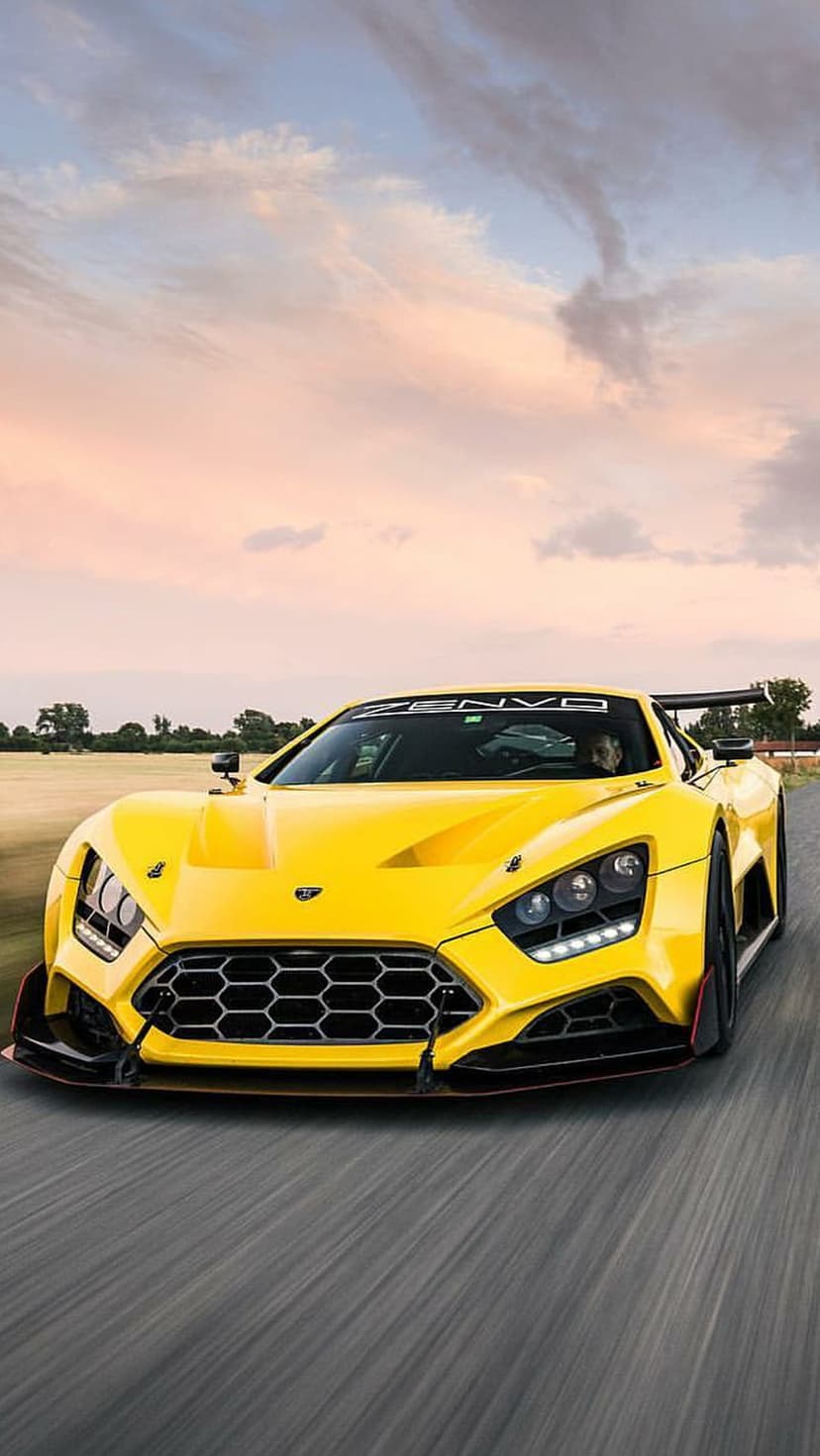 Yellow Zenvo ST1 supercar driving on a road under a pink sunset sky with motion blur and glowing LED headlights.