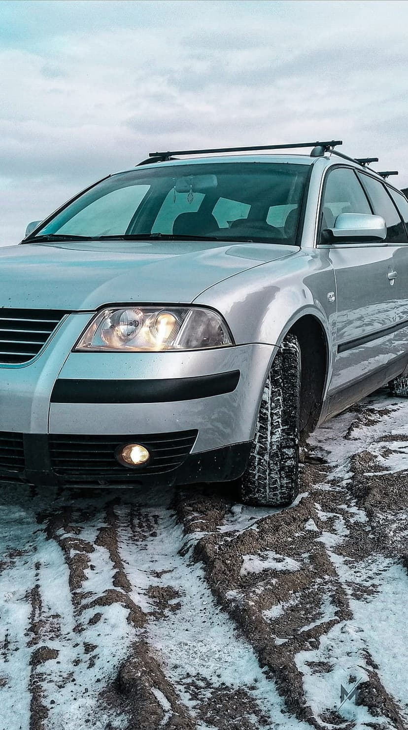 Silver Volkswagen Passat station wagon with glowing headlights parked on a muddy, snow-covered dirt road at dusk.