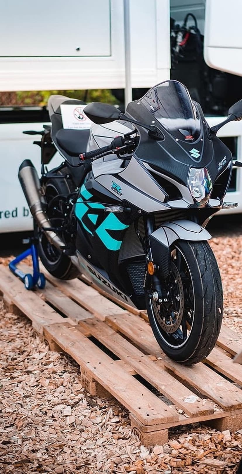 Suzuki GSX-R sportbike with teal accents sits on wooden pallets over a bed of wood chips in an outdoor setting.