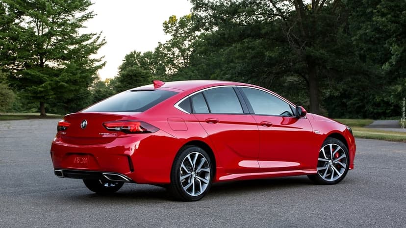 Red Buick Regal GS sport sedan on asphalt with a lush forest background and soft natural evening lighting.