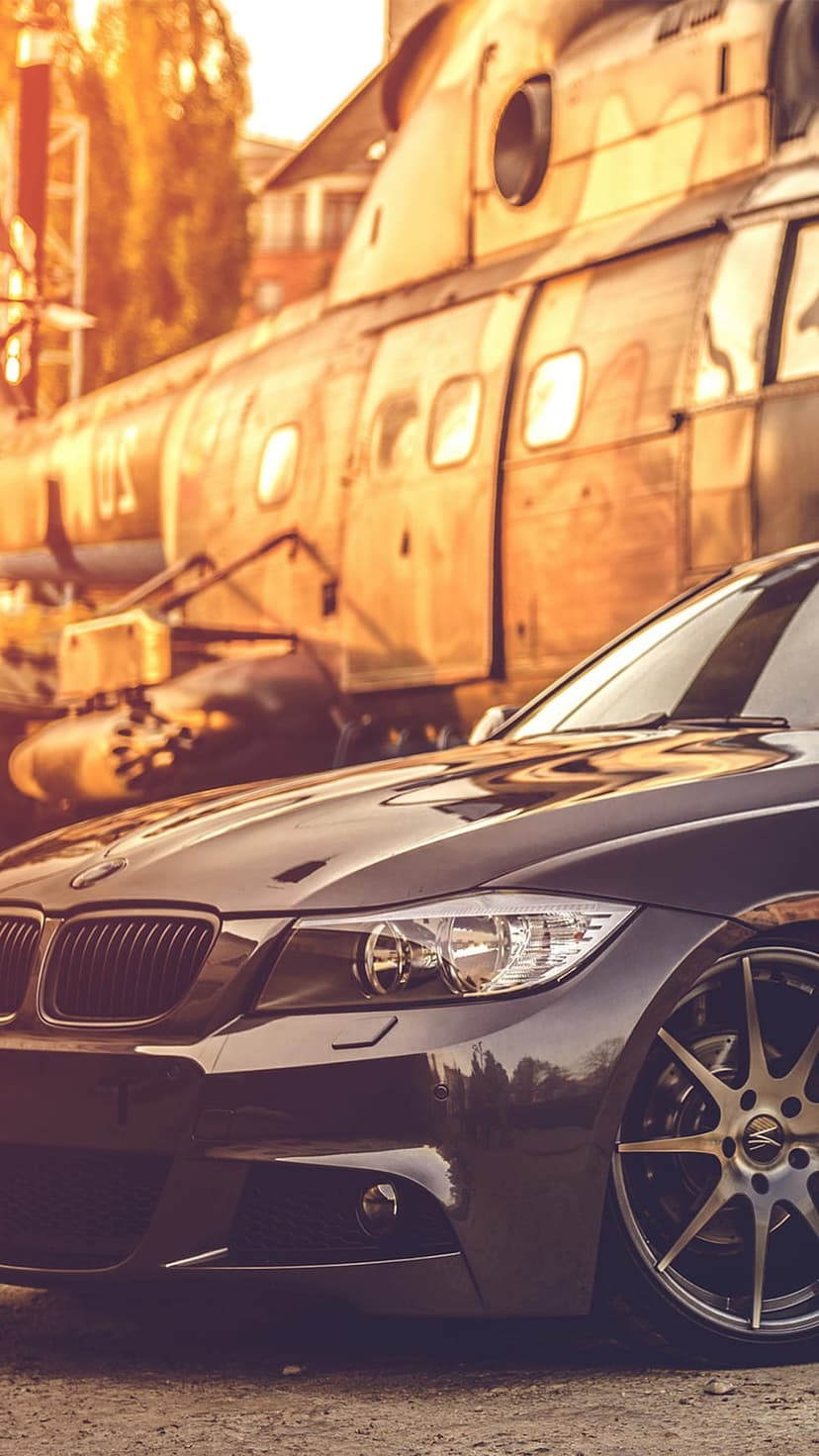 Black BMW car parked beside a large weathered vintage helicopter during a dramatic golden hour sunset outdoors.
