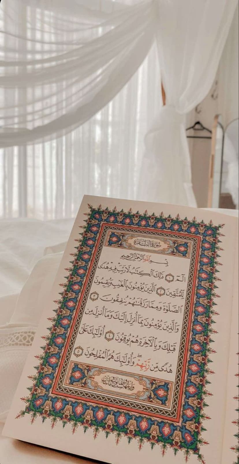 Open Quran with ornate borders sits on white bedding next to flowing sheer curtains in soft natural light.
