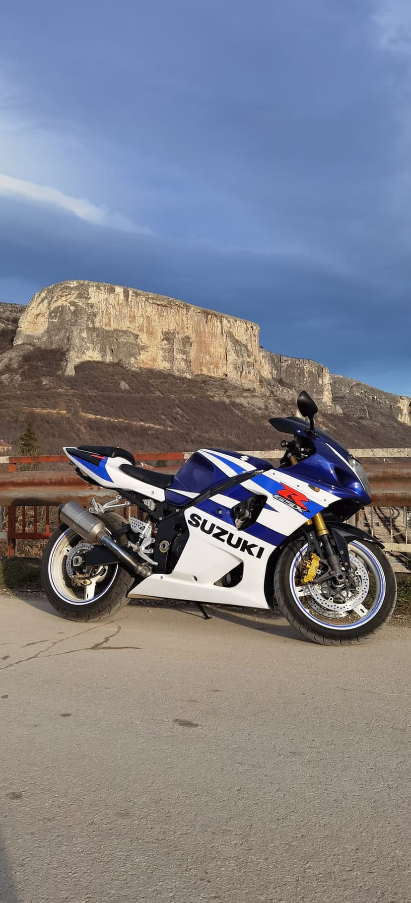 Blue and white Suzuki motorcycle parked on an asphalt road with rugged, rocky mountain peaks in the background.