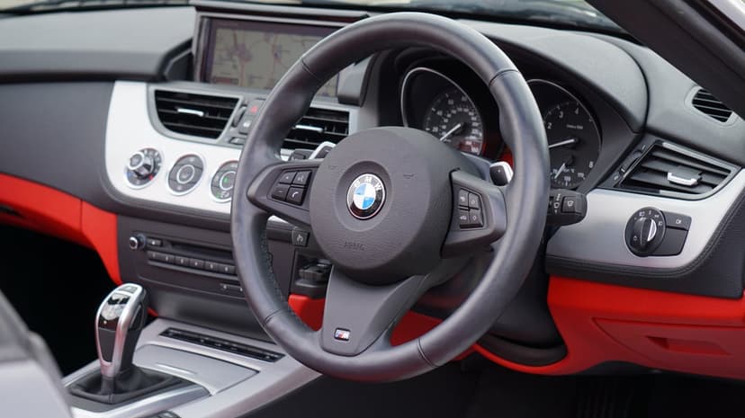 BMW leather steering wheel with red stitching and logo in a car cockpit with dashboard and gear shift.
