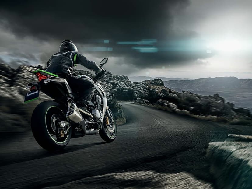 Motorcyclist on a Kawasaki sportbike leaning into a mountain road curve under a dramatic stormy sky.