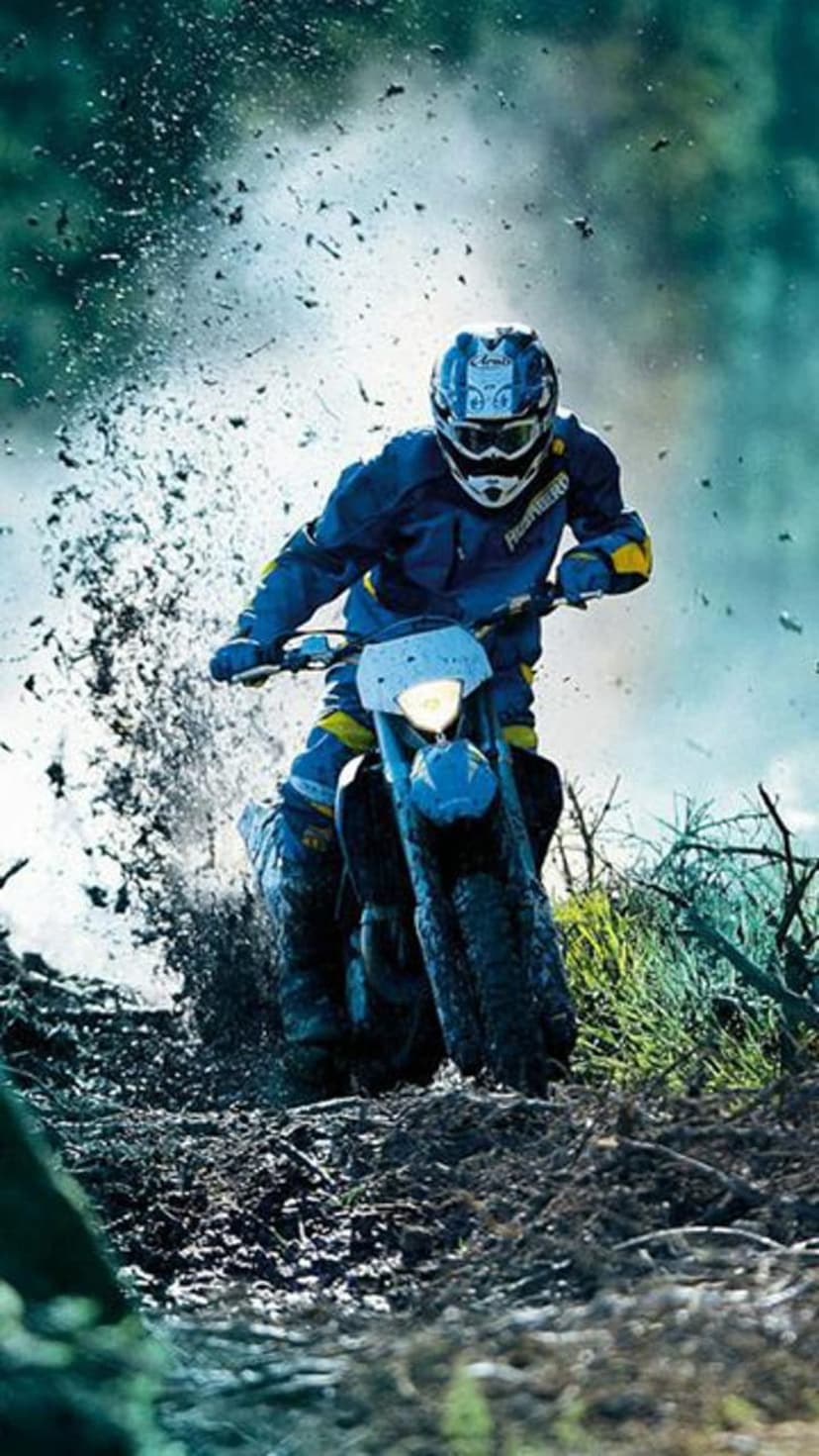 Motocross rider in blue and yellow gear leans into a turn while splashing through mud and water on a dirt track.