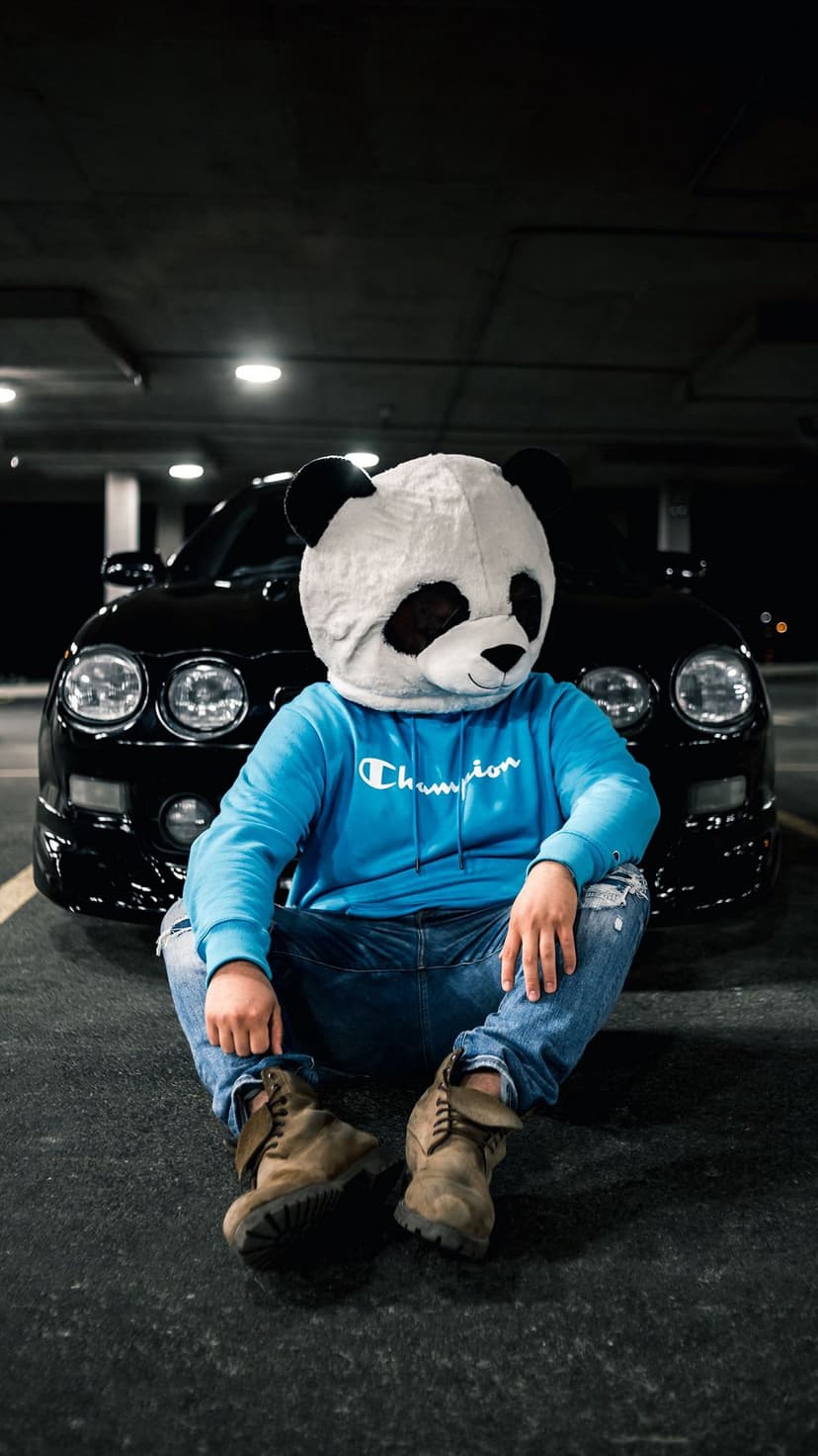 Person wearing a panda mask and blue hoodie sits by a black sports car in a dark underground parking garage.