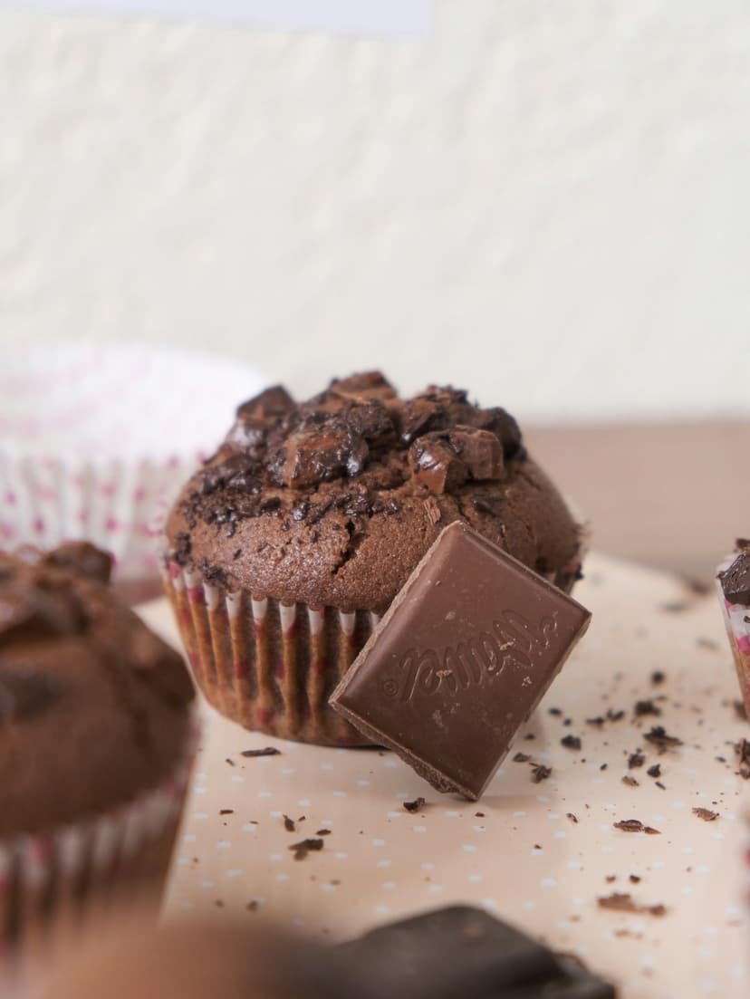 Dark chocolate muffins topped with melting cocoa chunks and a chocolate bar piece in warm, moody bakery lighting.