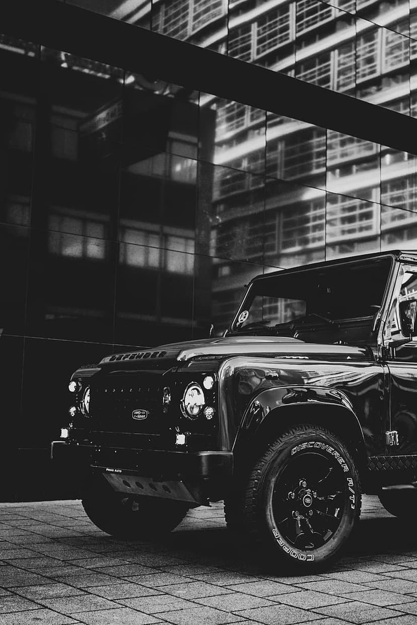 Black Land Rover Defender SUV parked on a reflective tiled surface near a modern glass building in monochrome.