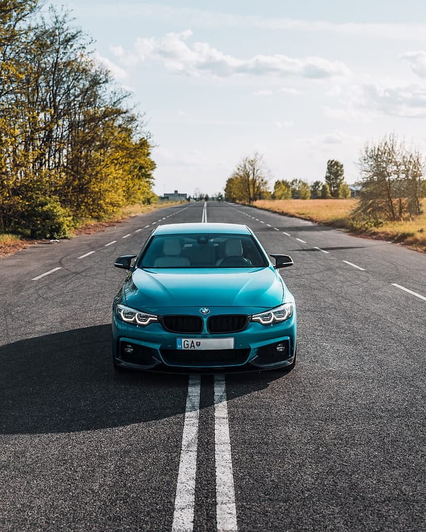 Teal BMW 4 Series coupe on an asphalt road with autumn trees and a bright sky in a low-angle front view.