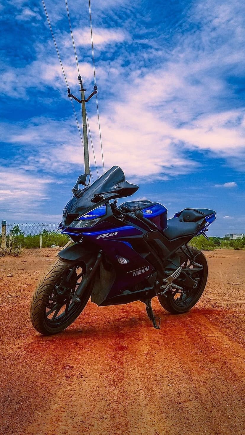Blue Yamaha R15 motorcycle on a dirt road under a cloudy sky with a utility pole in the background scenery.