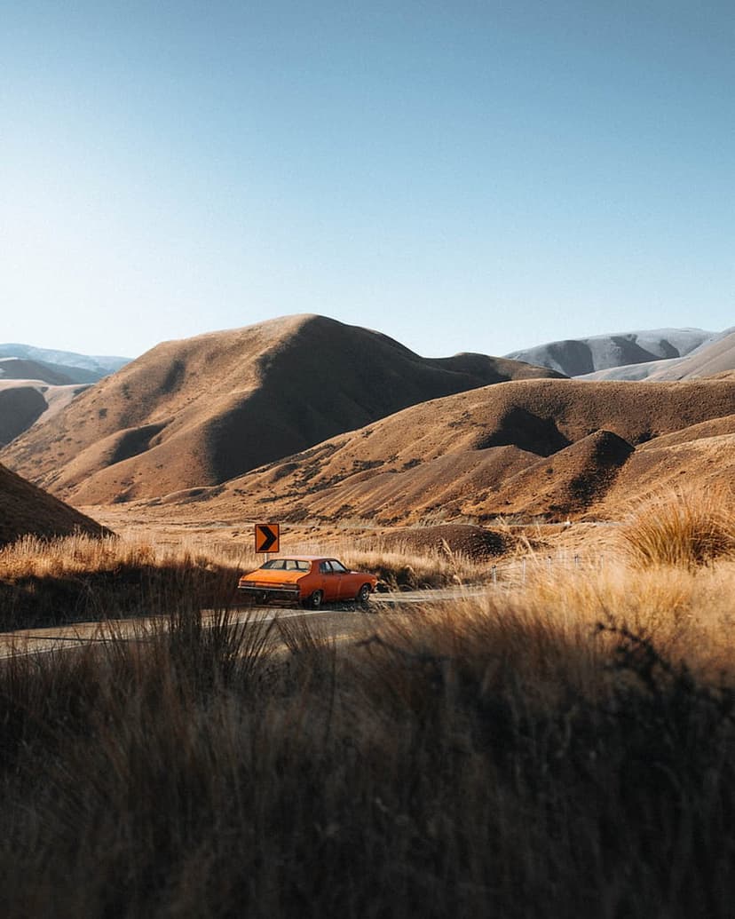 Orange vintage sedan navigates a curving asphalt road amidst rolling golden hills and distant rugged mountains.