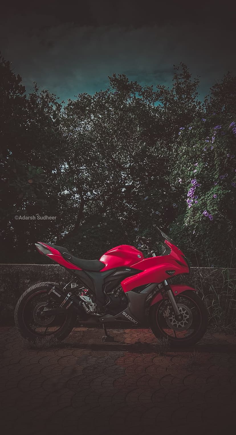 Red Suzuki sportbike parked on asphalt surrounded by green trees and purple flowers under a dramatic moody sky.