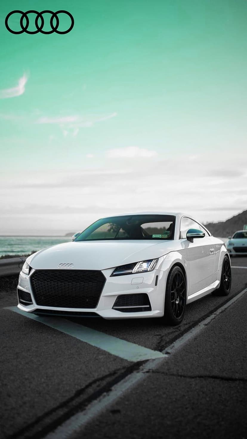 White Audi TT sports car on a seaside road with large Audi rings floating in a turquoise sky over the ocean.