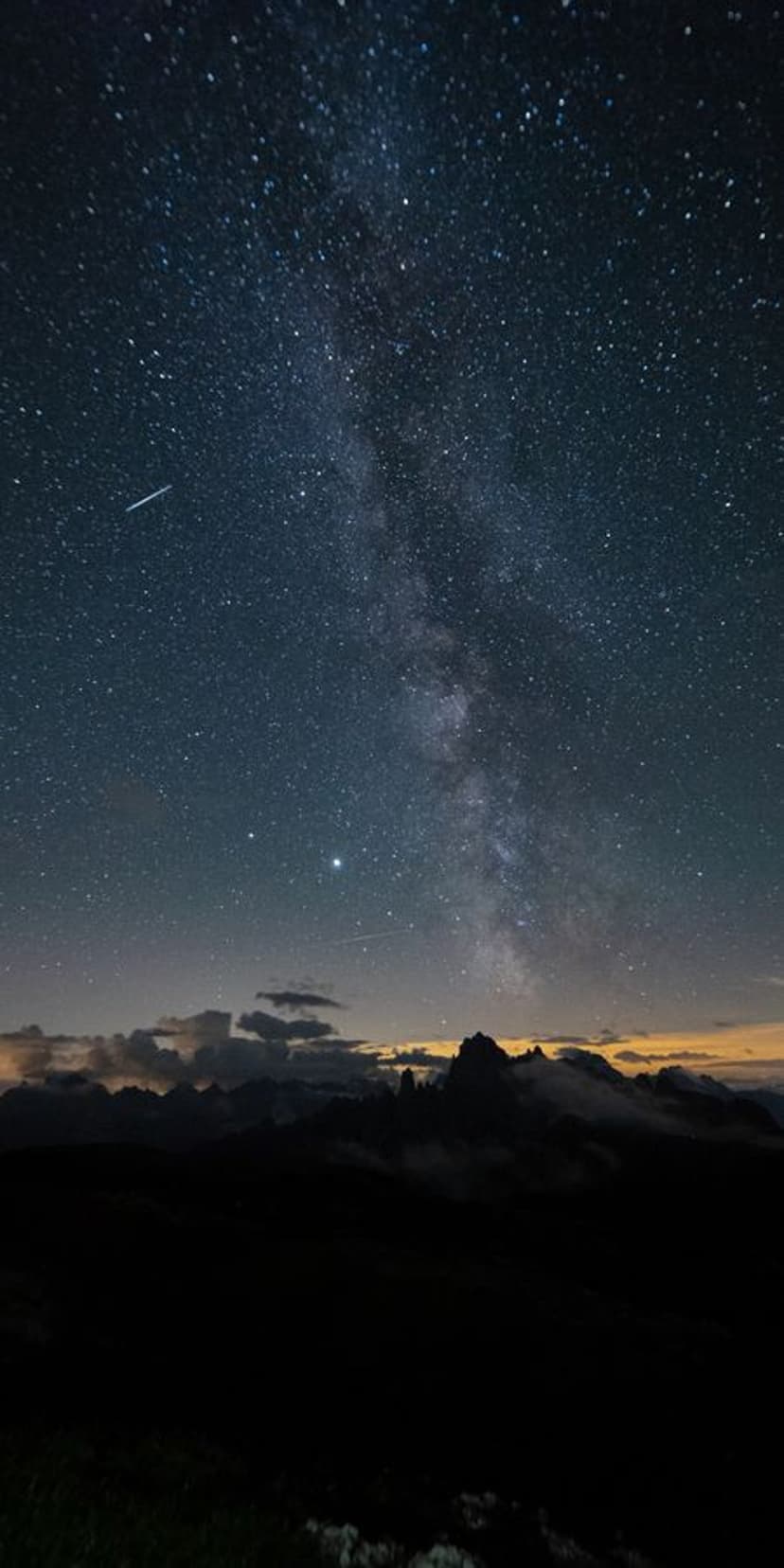 Jagged mountain peaks silhouetted against a purple Milky Way galaxy with a visible shooting star and low clouds.