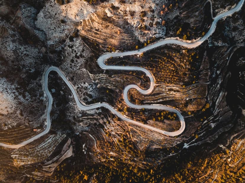 Serpentine asphalt road weaves through rugged mountains as golden sunset light creates long shadows on the rocks.