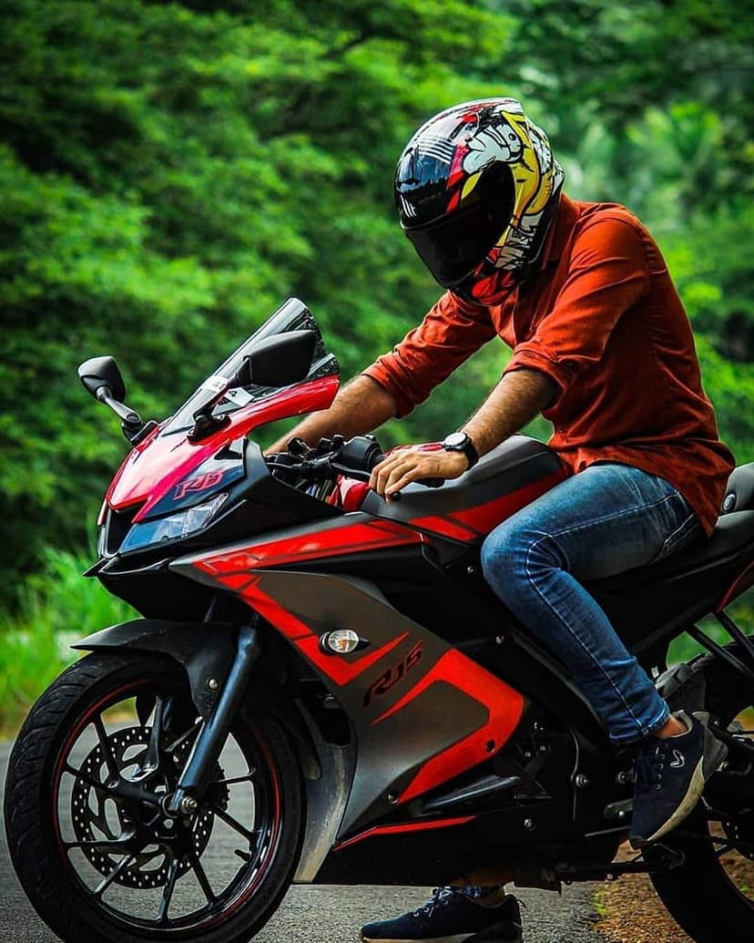 Man in a red shirt and helmet sits on a red sport motorcycle on an asphalt road with green trees behind him.