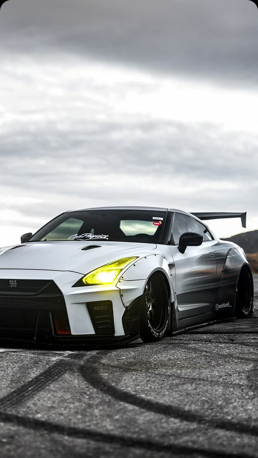 White Nissan GTR with a wide body kit, yellow headlights, and rear spoiler parked on asphalt with tire marks.