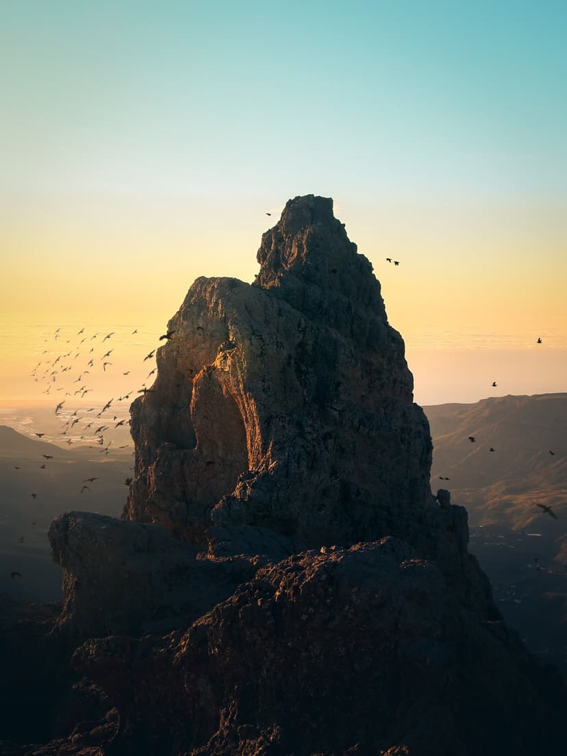 Birds fly around a sharp mountain summit under a glowing sunset sky with deep orange and purple gradients.