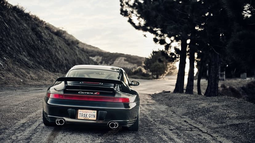 Black Porsche 911 Carrera 4S parked on a scenic winding road against a backdrop of hills and dark trees.