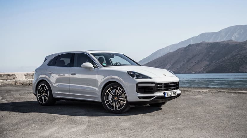 White Porsche Cayenne luxury SUV parked on a coastal road with blue water and rugged mountains under a clear sky.