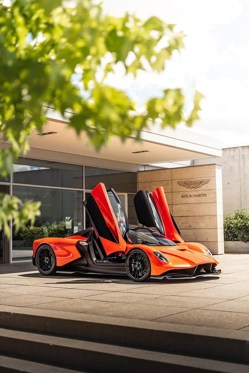 Orange Aston Martin Valkyrie Spider parked with dihedral doors open outside a modern building in bright sunlight.