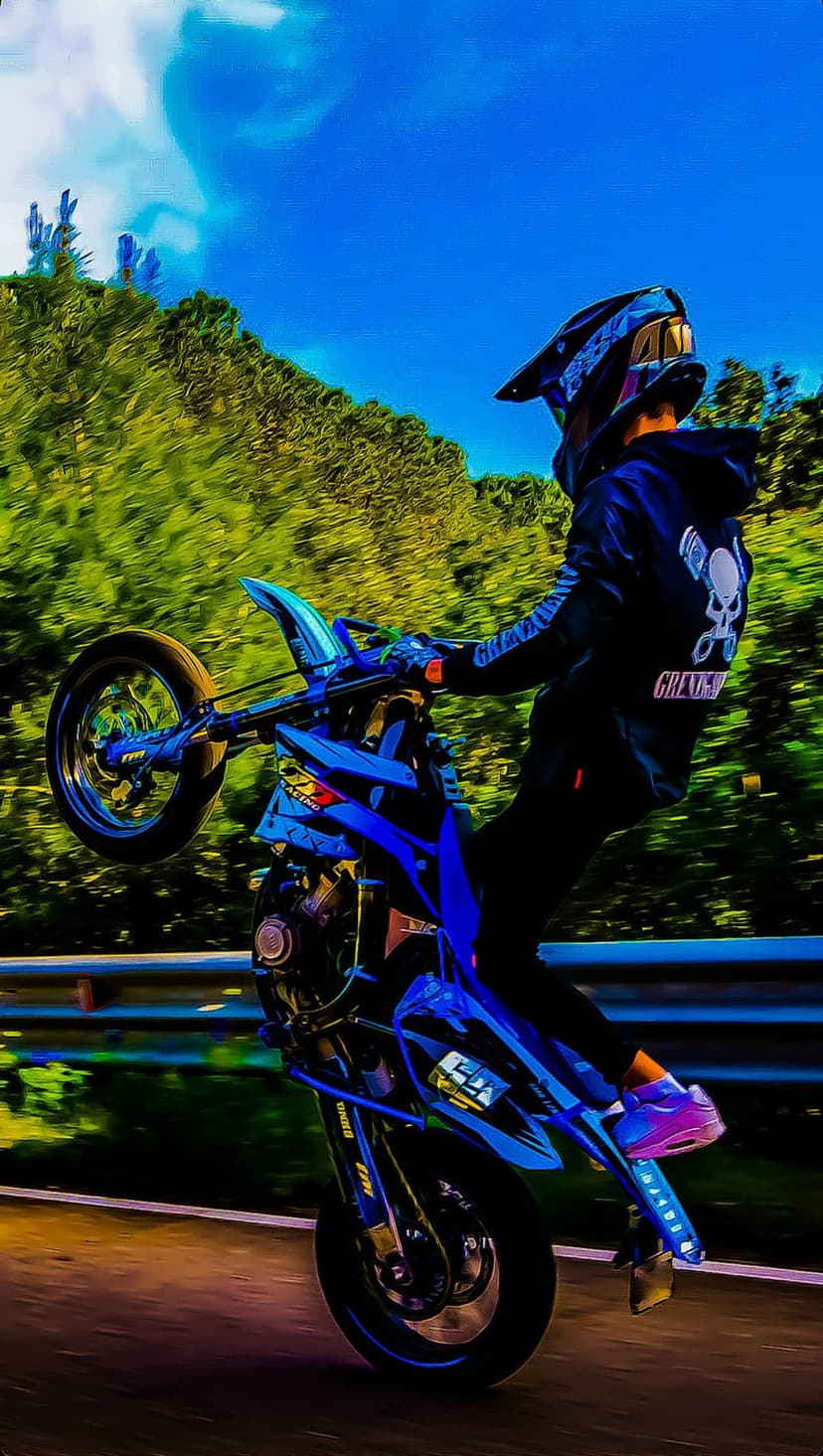 Motorcyclist on a blue sportbike performs a wheelie with the front wheel raised against a blurred green forest.