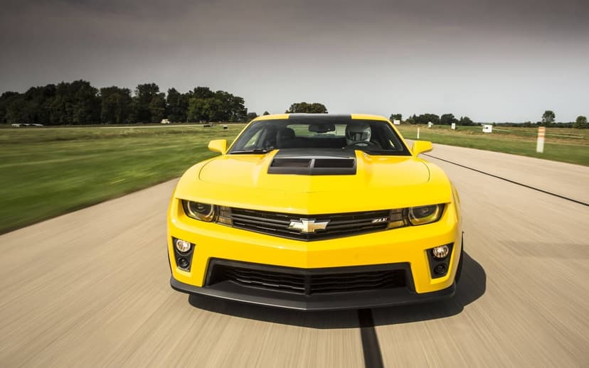 Front view of a yellow Chevrolet Camaro ZL1 speeding on a track with motion blur and a helmeted driver.