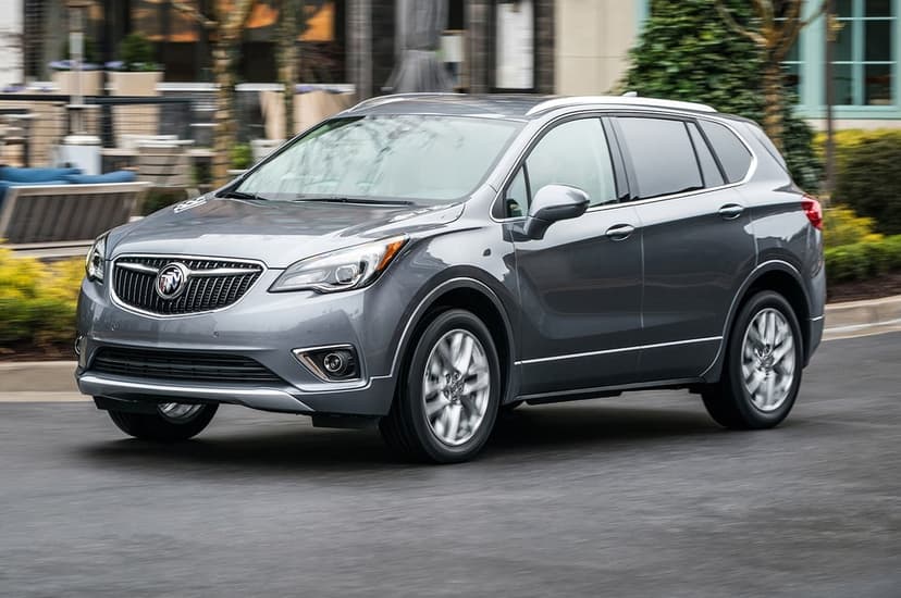 Grey Buick Envision SUV driving on a city road with blurred urban buildings and green trees in the background.