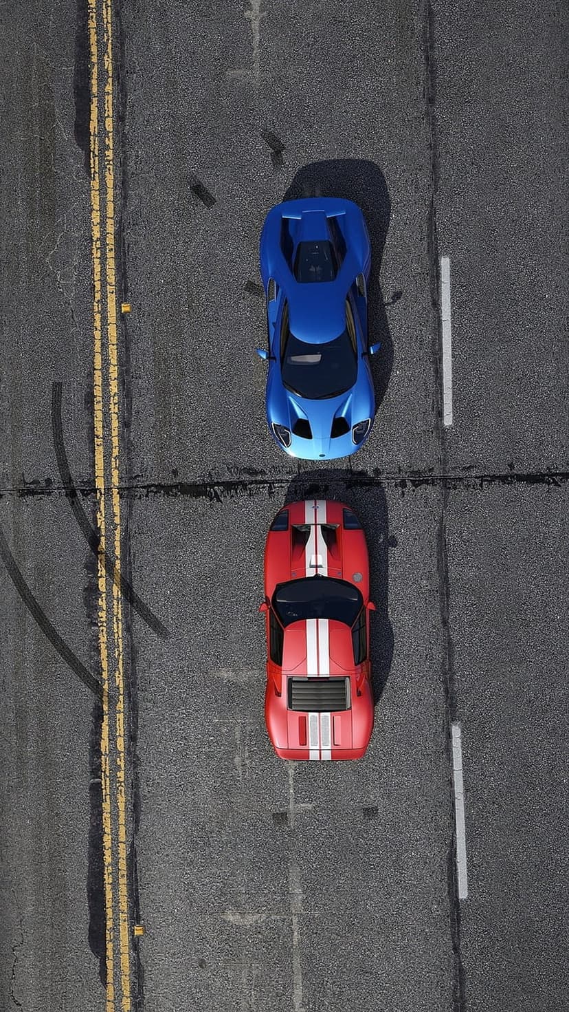 Red and blue sports cars race side-by-side on a textured asphalt road from a direct overhead aerial viewpoint.