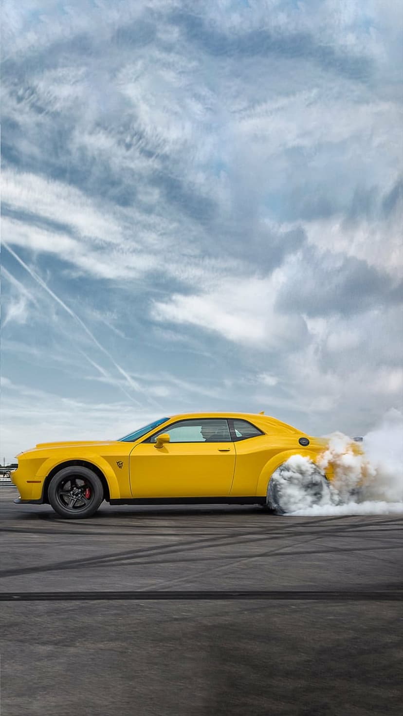 Yellow Dodge Challenger muscle car drifting on asphalt while emitting thick white smoke from spinning rear tires.