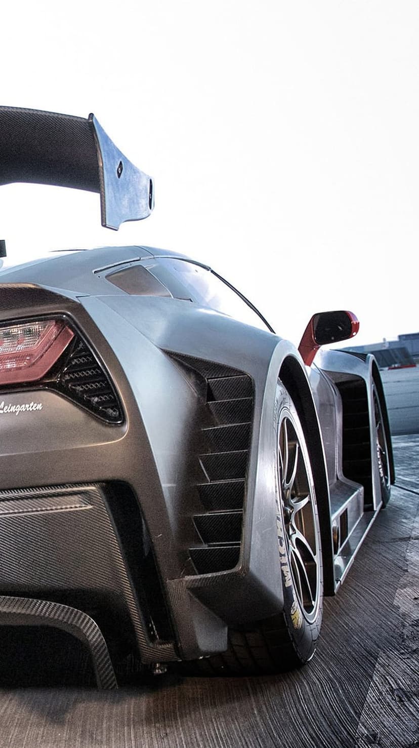 Rear view of a matte black race car with visible carbon fiber textures and an aerodynamic wing against a gray sky.
