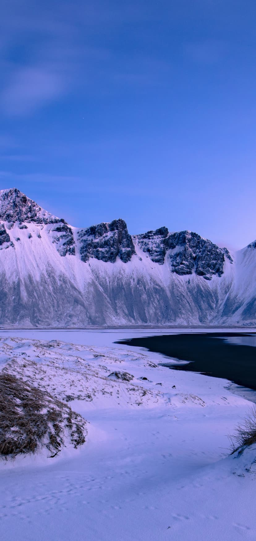Snow-covered Vestrahorn mountain peaks overlooking icy black sand dunes and frozen beach water in Iceland.