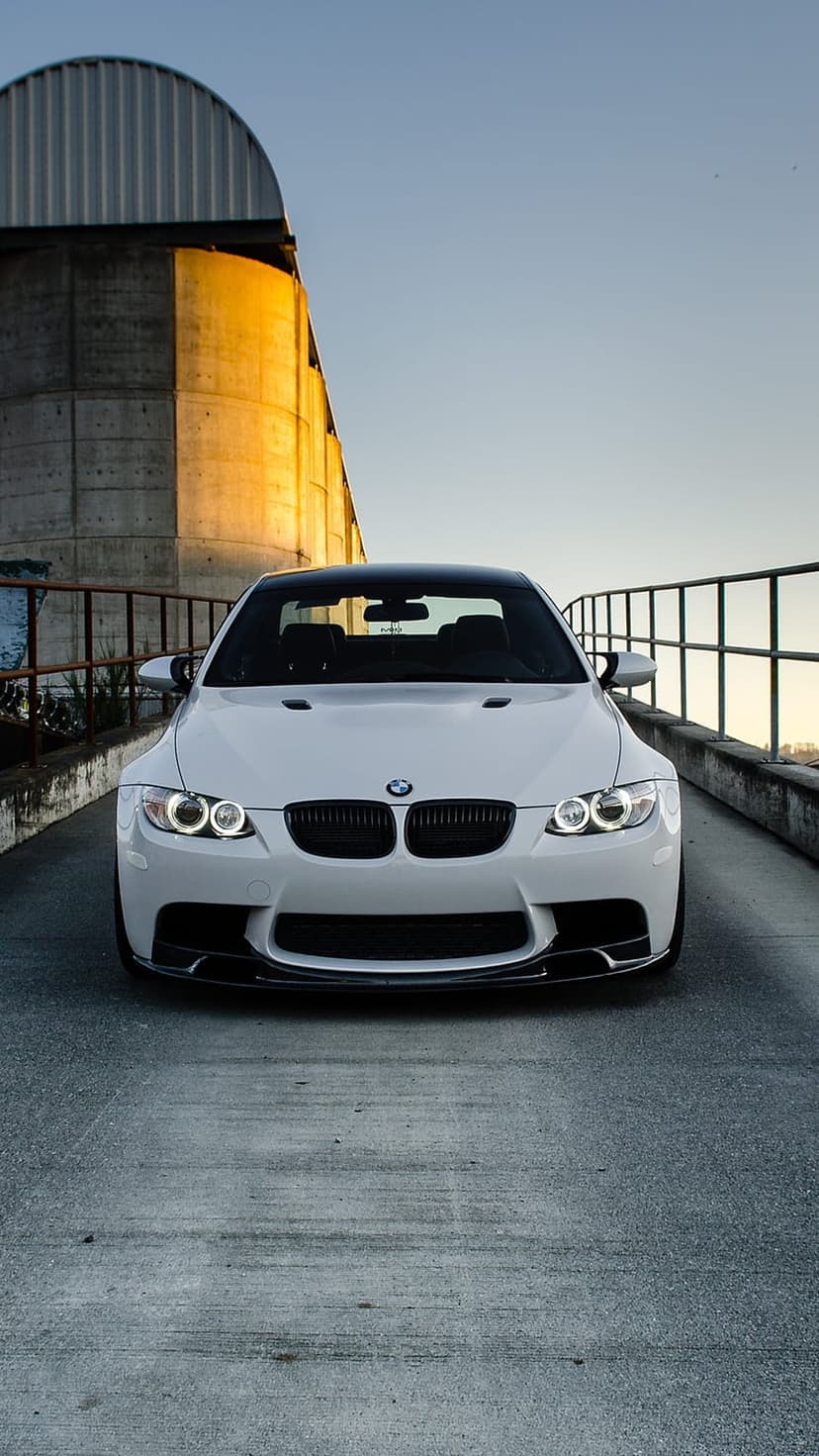 White BMW M3 parked on concrete beside an industrial silo under a golden sunset with clear blue skies.