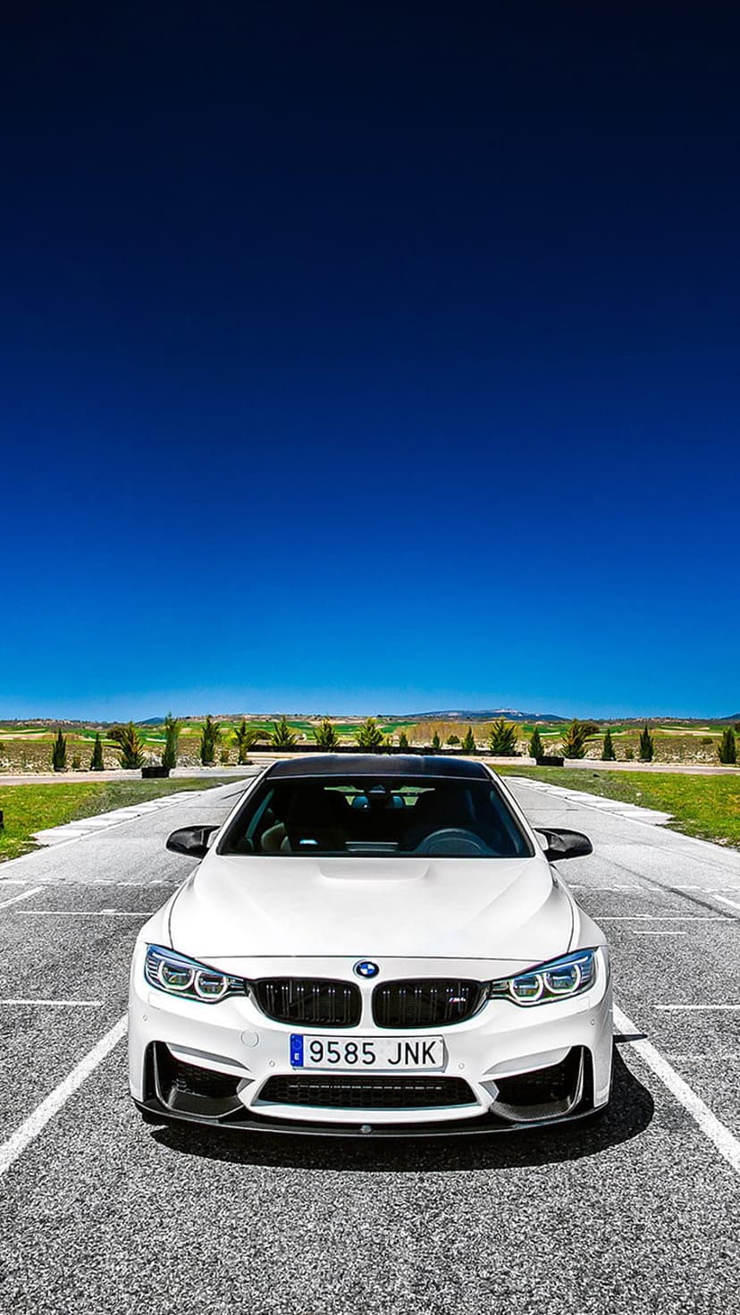 White BMW M4 with headlights on parked on a race track with green hills and a blue sky in the background.
