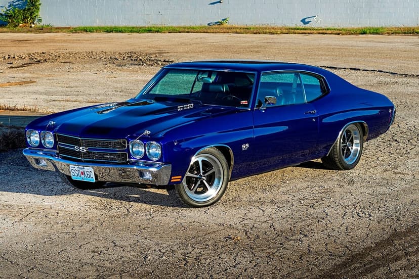 Blue 1970 Chevrolet Chevelle SS muscle car with black hood stripes parked on sunlit asphalt by a brick wall.