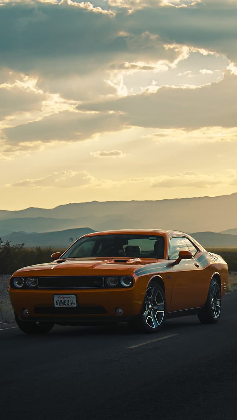 Orange Dodge Challenger parked on an asphalt road against a backdrop of purple mountains and a sunset sky.