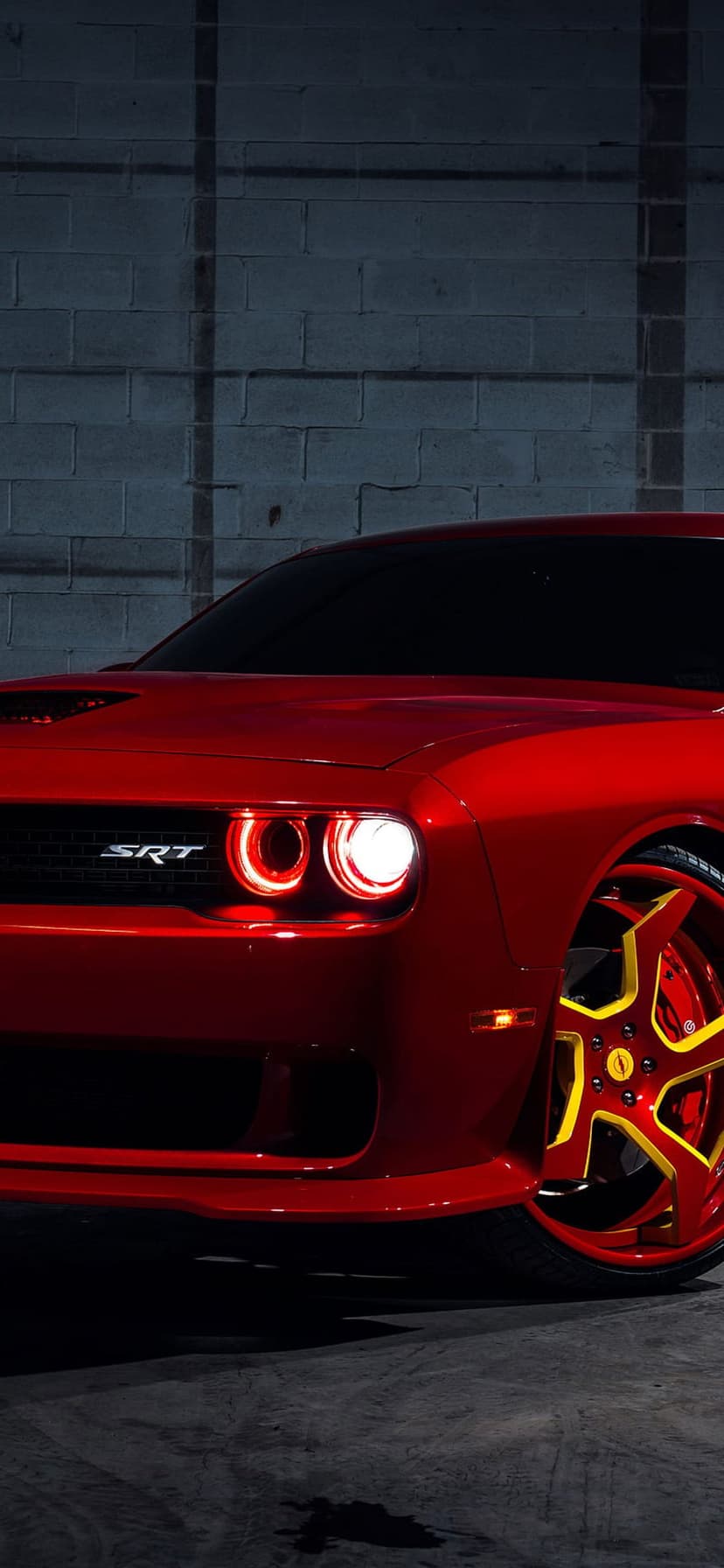Red Dodge Challenger SRT front end with yellow rims and glowing halo headlights against a brick wall.