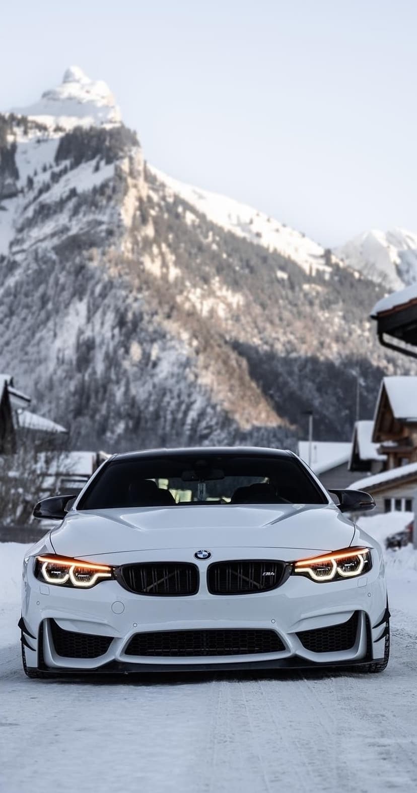 White BMW M4 with illuminated headlights parked on a snowy road against a backdrop of steep alpine mountains.