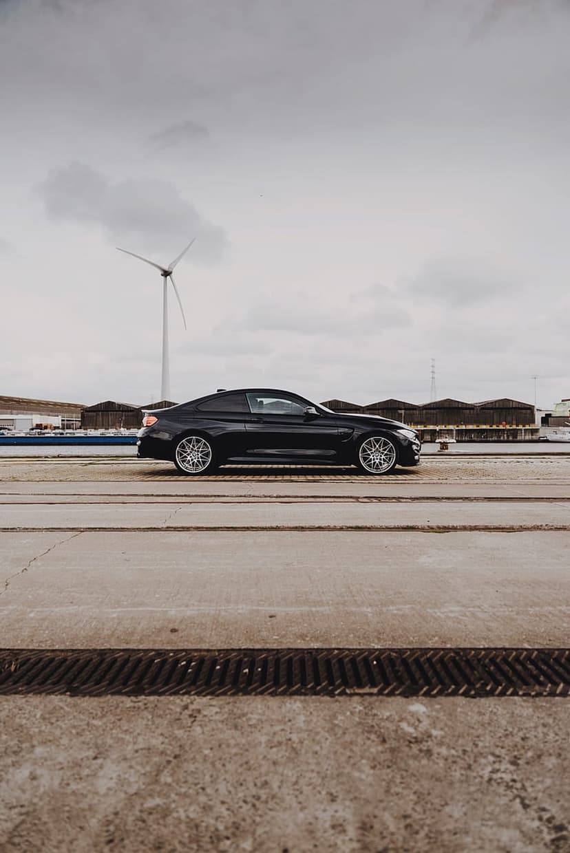 Black BMW M4 sports car parked on asphalt with a large wind turbine under a cloudy sky in the background.