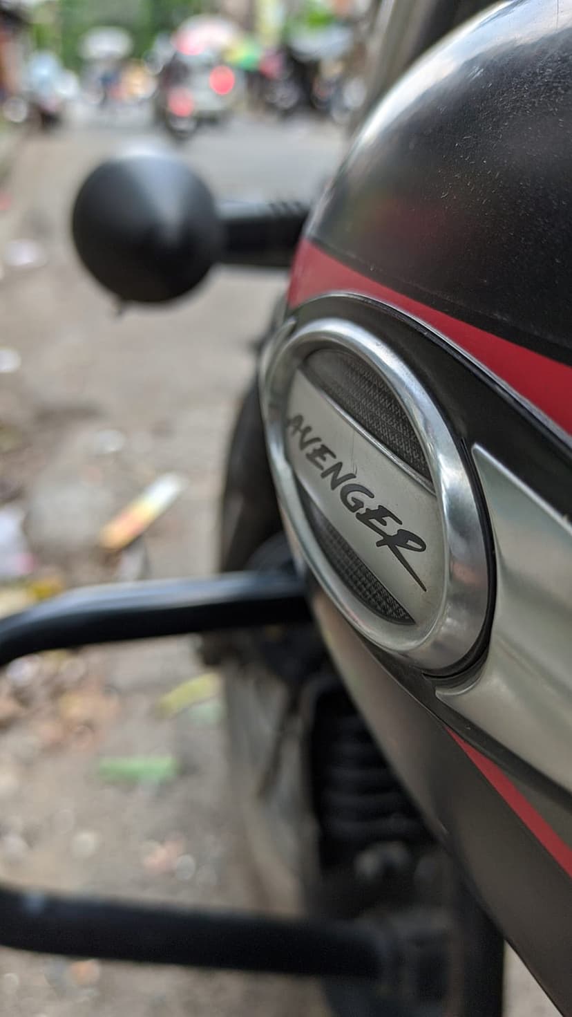 Avenger motorcycle logo on a brushed metal fuel tank with red accents and a blurred urban street background.