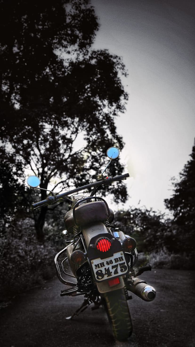 Royal Enfield motorcycle rear view on a dusty forest road with a glowing red taillight and dark tree silhouettes.