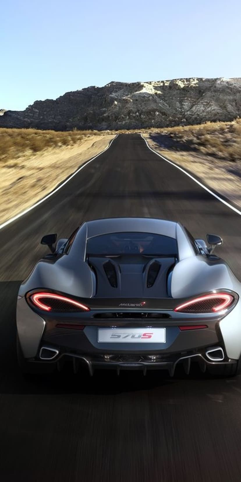 McLaren 570S rear view racing on an asphalt road through a desert with mountains under a clear blue sky.