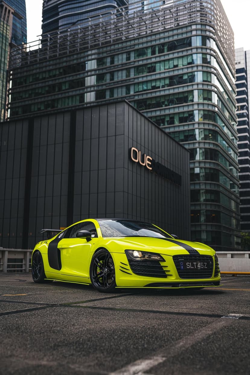 Neon yellow Audi R8 sports car with black racing stripes parked in front of a sleek glass skyscraper at night.