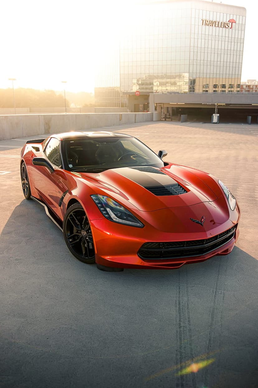 Orange Corvette C7 parked on a concrete rooftop at sunset with a tall glass skyscraper reflecting golden light.