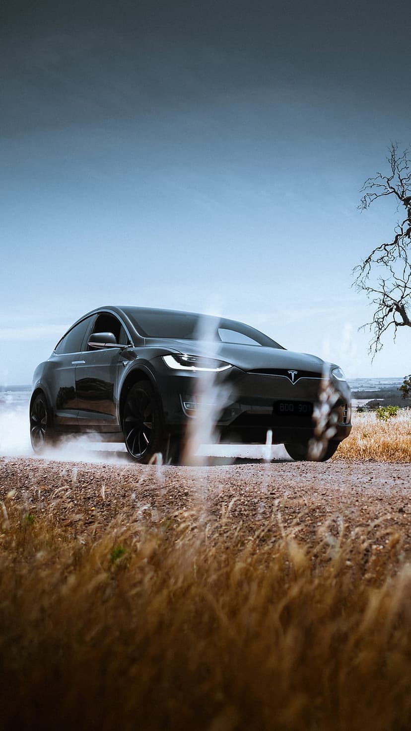 Black Tesla Model X SUV drifting on a dusty dirt road with glowing headlights under a hazy blue sky.